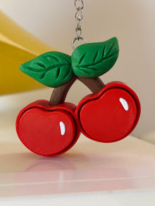 Vista frontale del fidget toy Portachiavi Ciliegie Clicker di Namii nei colori rosso, marrone e verde, immortalato appeso mentre tocca leggermente la superficie con una delle due ciliegie stampate in 3D.