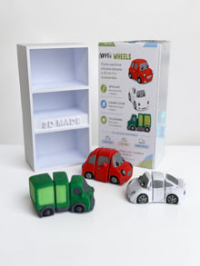 Inquadratura frontale che mostra due confezioni di Namii Wheels, una esposta sul davanti e una sul retro, caratterizzate da una scatolina a tre scomparti; davanti al packaging sono posizionati i tre veicoli snodati della collezione: il camioncino verde, la spider bianca e la macchinina rossa.